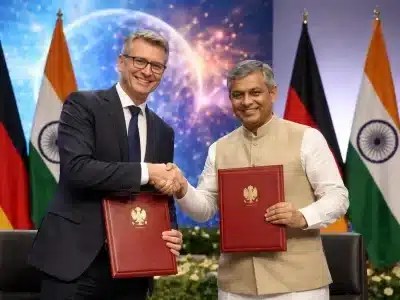 Zwei Minister schütteln sich mit Vertragsmappen die Hände. Im Hintergrund stehen die Flaggen von Deutschland und Indien