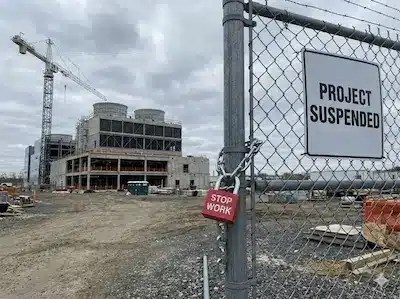 Baustelle mit unfertigem Industriegebäude hinter Zaun; Schild „Project Suspended“ und Schloss mit „Stop Work“.