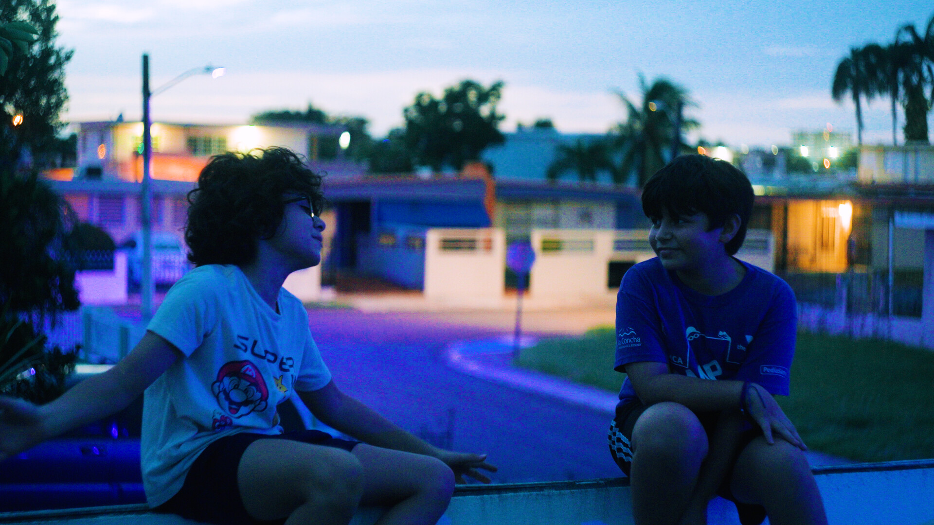 Two children sit on a low wall at dusk in a residential street, streetlights and houses glowing behind them.
