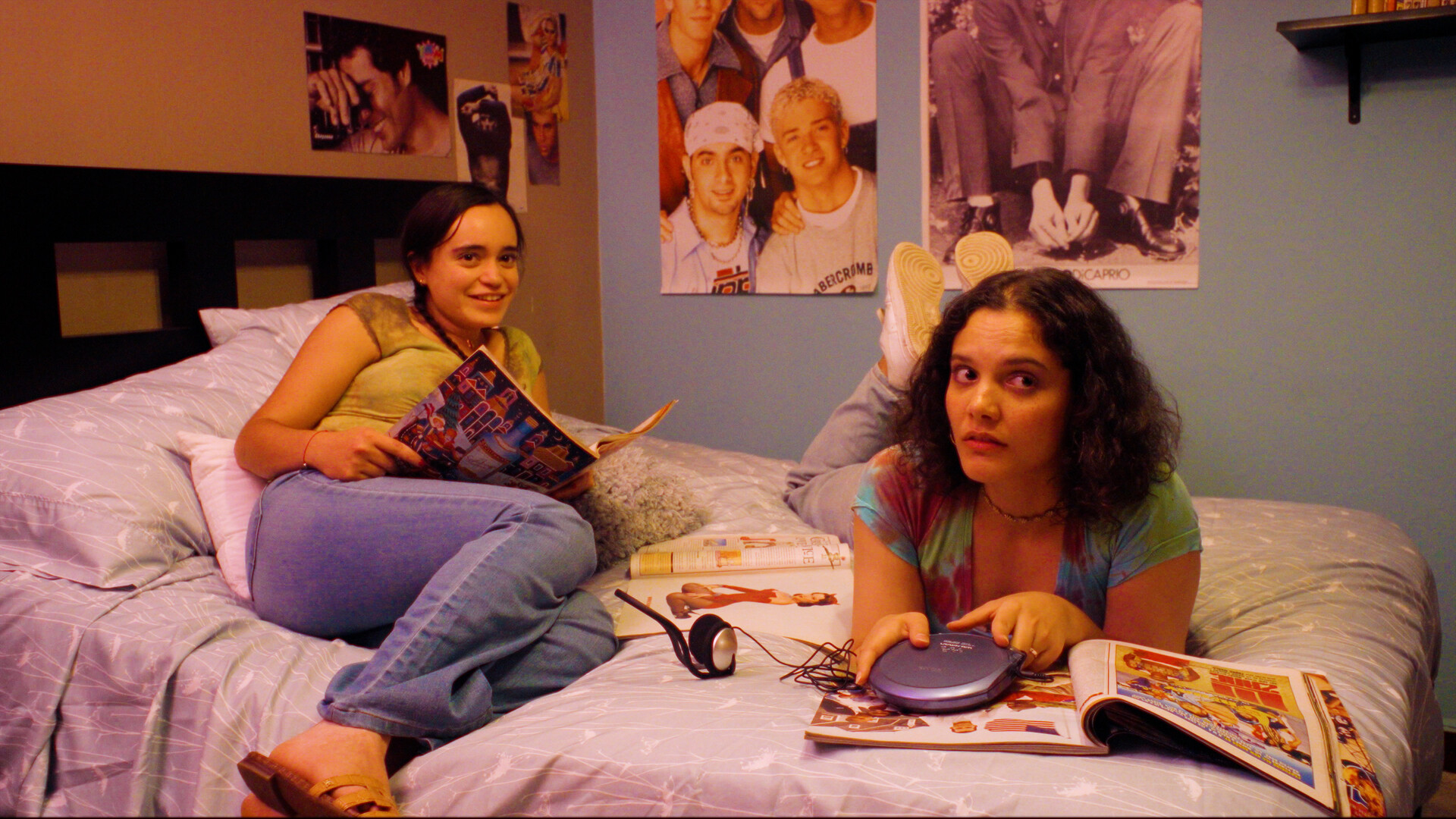 Two teenage girls lie on a bed surrounded by magazines and a portable CD player, band posters on the wall behind them.