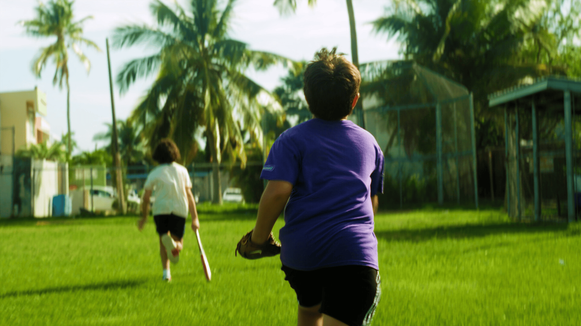 Two boys run across a grass field lined with palm trees, one carrying a bat and one wearing a baseball glove.
