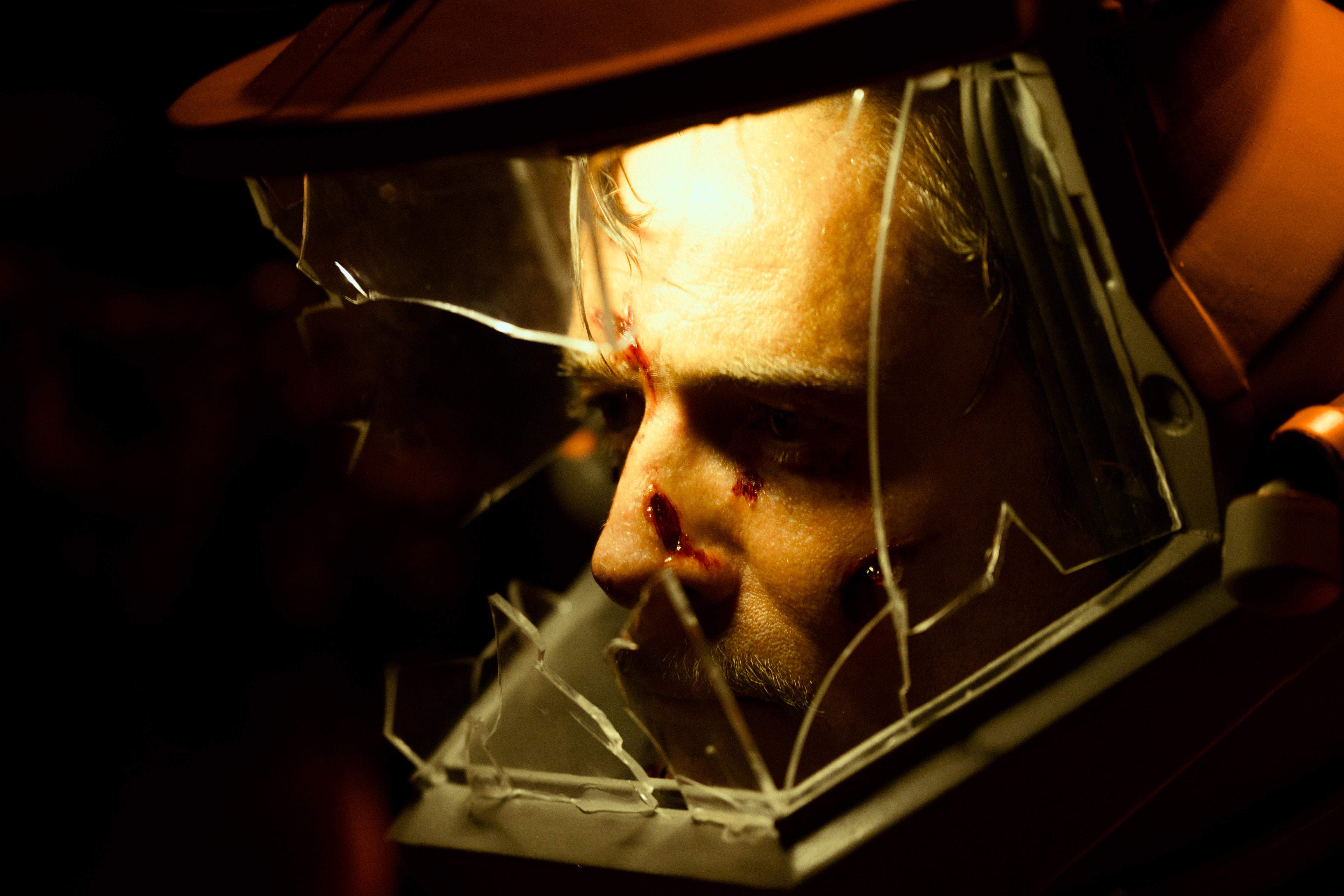 Close-up of a man's face lit in orange behind a shattered glass visor, with cuts on his forehead and nose, in a still from Crawlers