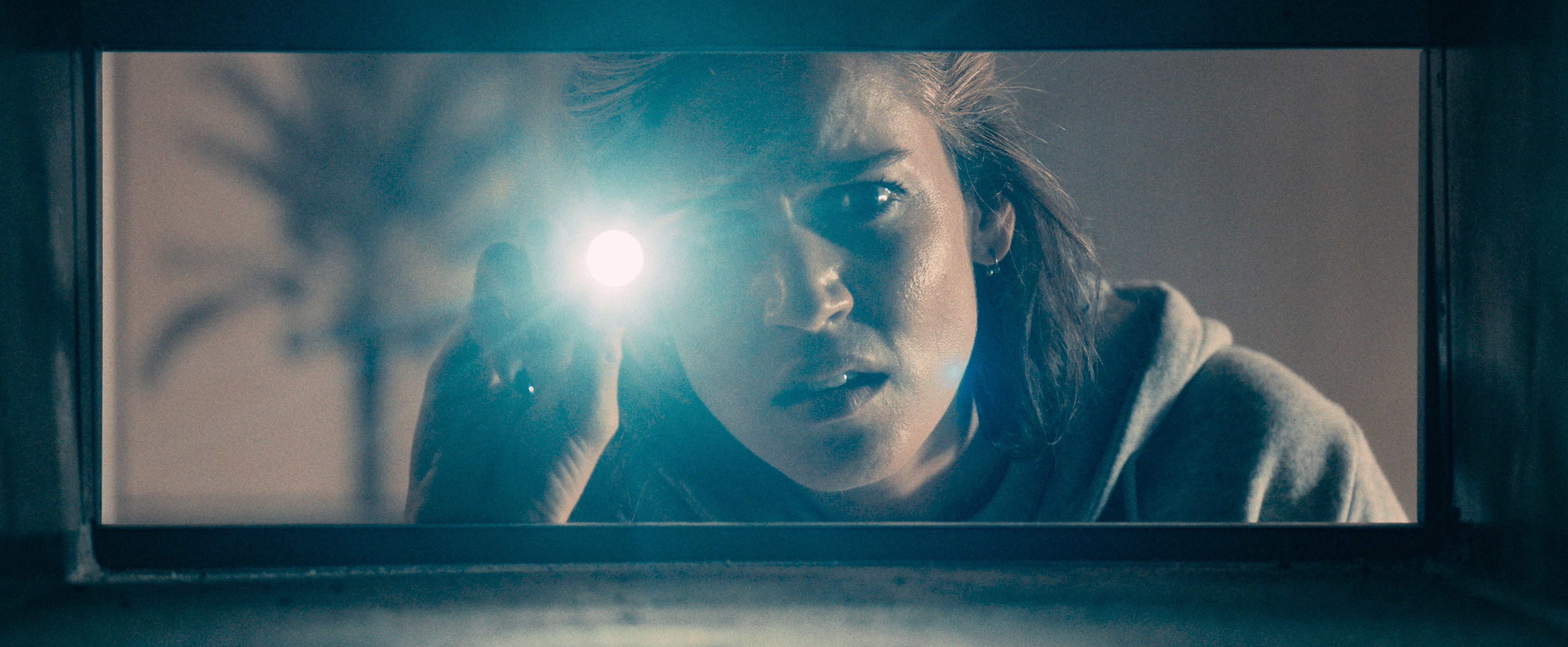 A woman in a gray hoodie points a bright flashlight toward the camera through a narrow opening in a still from Crawlers