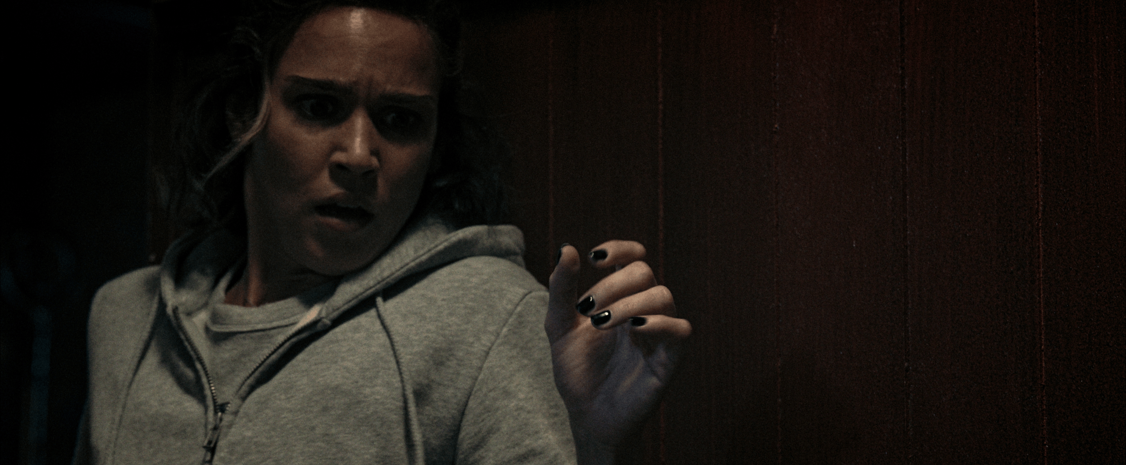A woman in a gray hoodie recoils in fear against a dark wood-paneled wall, one hand raised with black nail polish, in a still from Crawlers