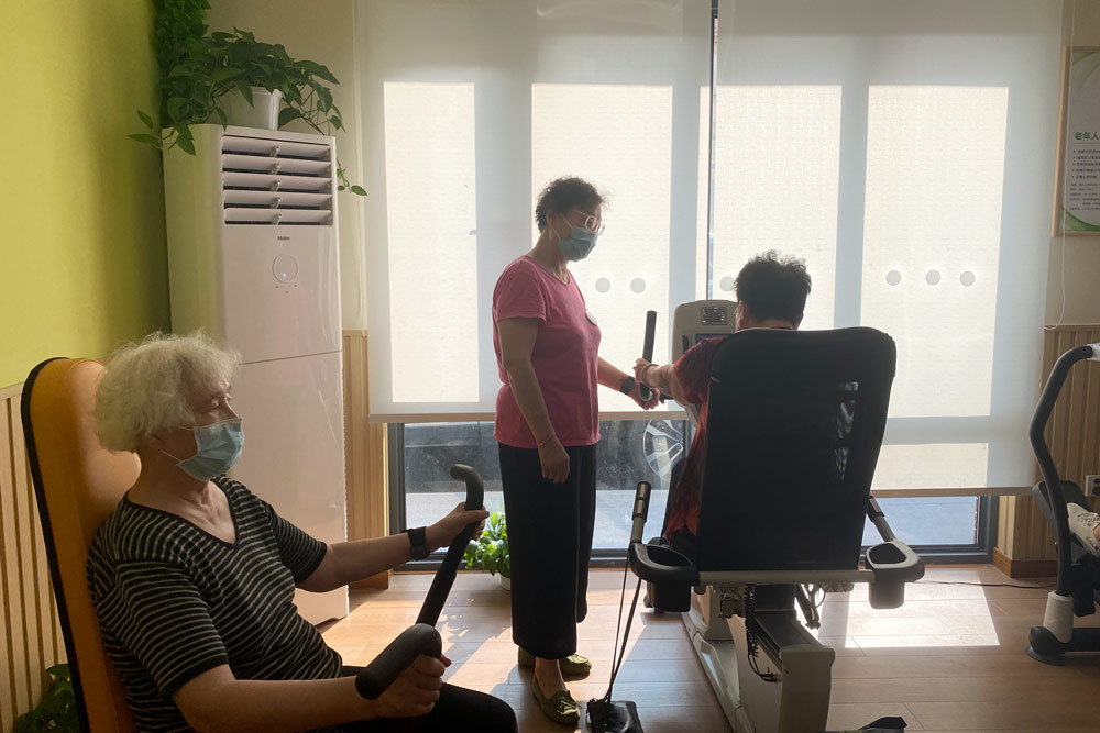 Pan Guoli (standing) shows an elderly resident how to use the machines at the gym where she works in Hongqiao Subdistrict, Shanghai, Aug. 19, 2021. Fan Yiying/Sixth Tone