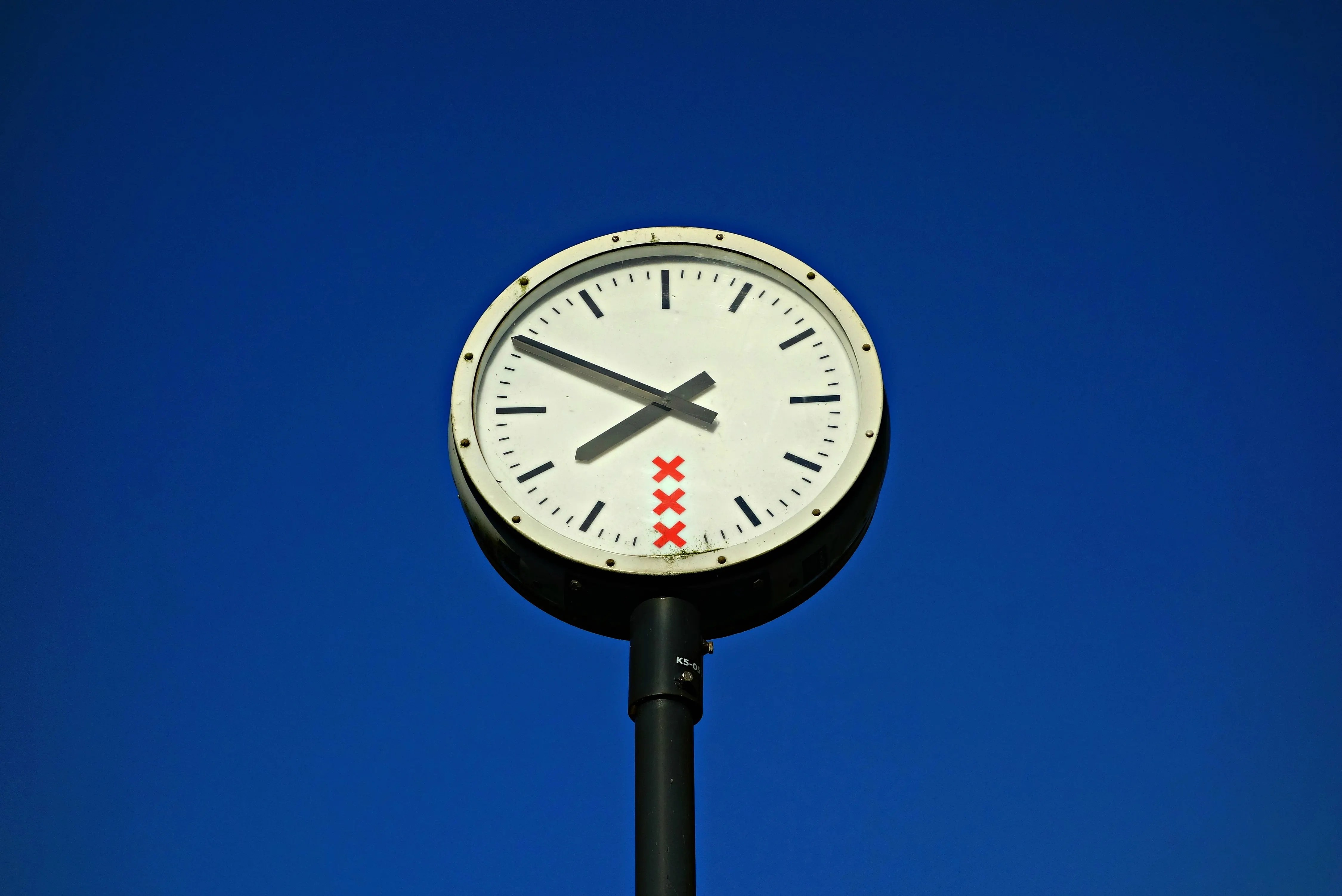 A public clock with red X's on its face stands against a clear blue sky.