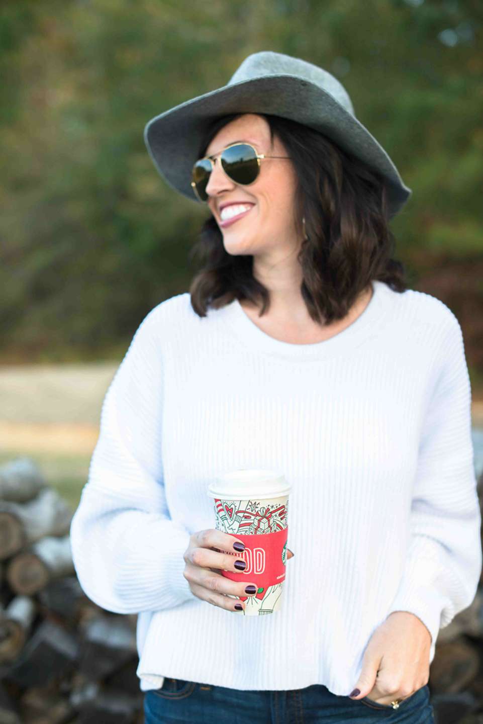 Smiling person in a gray wide-brimmed hat and white sweater holds a festive takeaway cup outdoors.