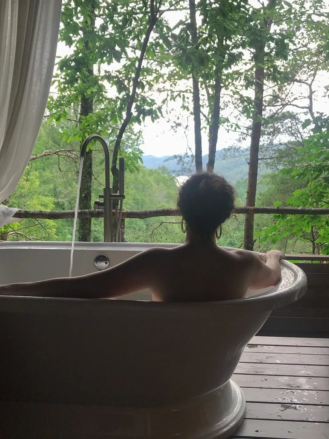 A person relaxes in an outdoor bathtub on a wooden deck, surrounded by lush greenery and trees. The scene is serene and idyllic, with a distant view of rolling hills.