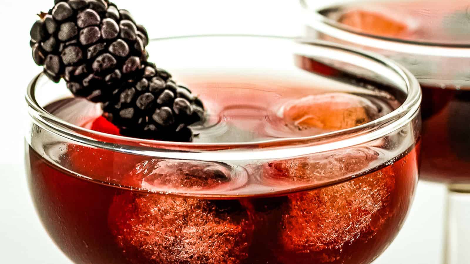 Two glasses of red cocktail with ice cubes, garnished with a blackberry on top, against a white background.