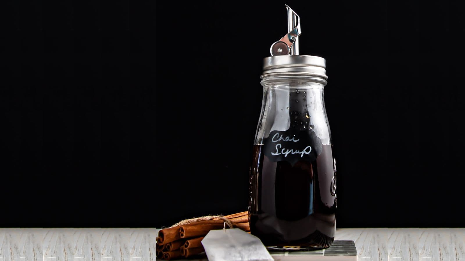 A glass jar labeled "chai syrup" filled with dark brown liquid, featuring a pour spout and a pen nib on top. it is placed on a white surface against a black background, with cinnamon sticks beside it.