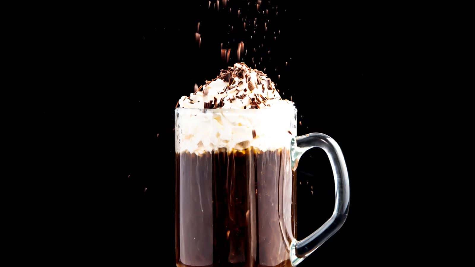 A glass mug of hot chocolate topped with whipped cream and chocolate shavings, with more shavings sprinkling down, set against a dark background.