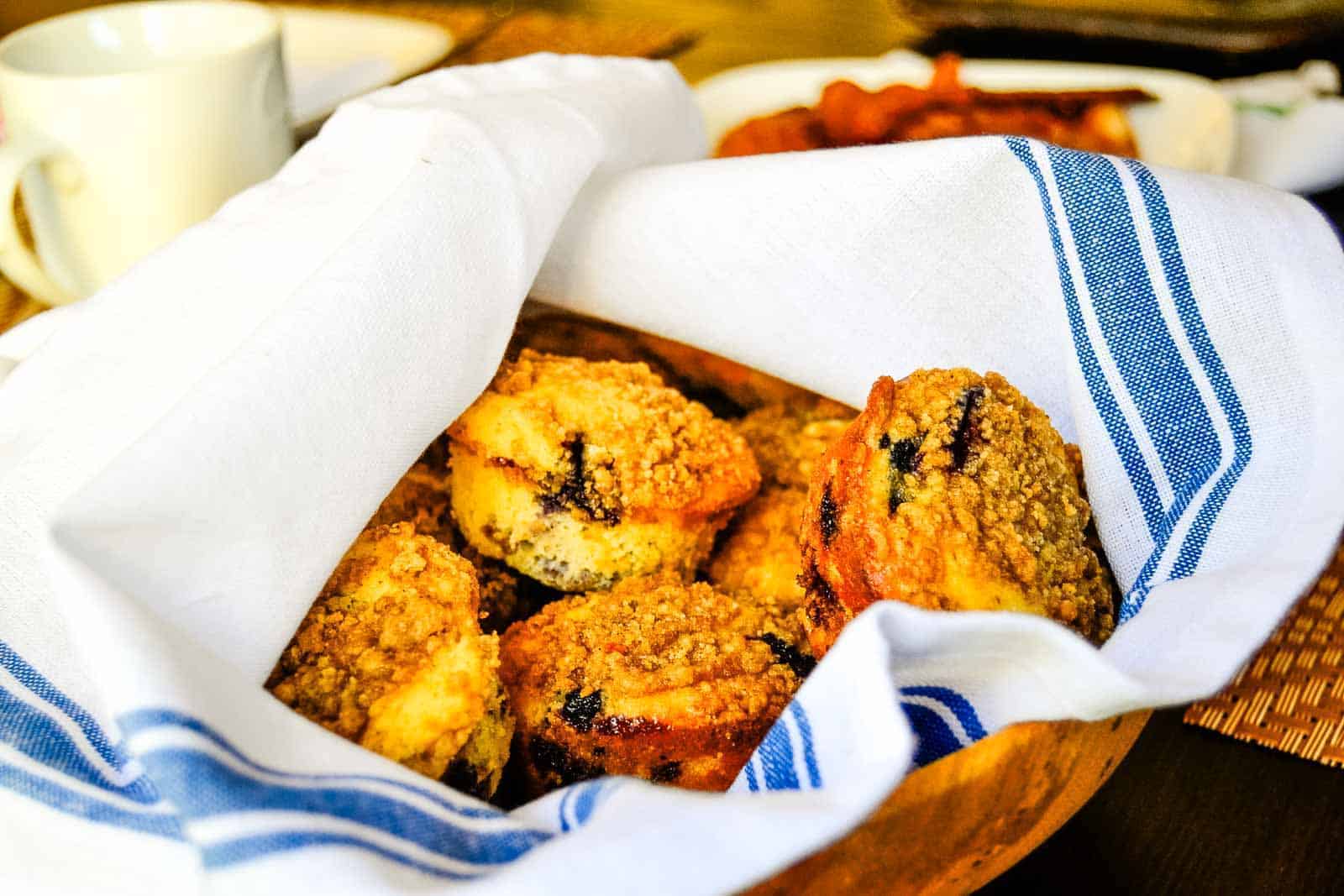 A wooden bowl lined with a white and blue striped cloth contains several streusel-topped breakfast breads. A white cup and other dishes are partially visible in the background on a table.