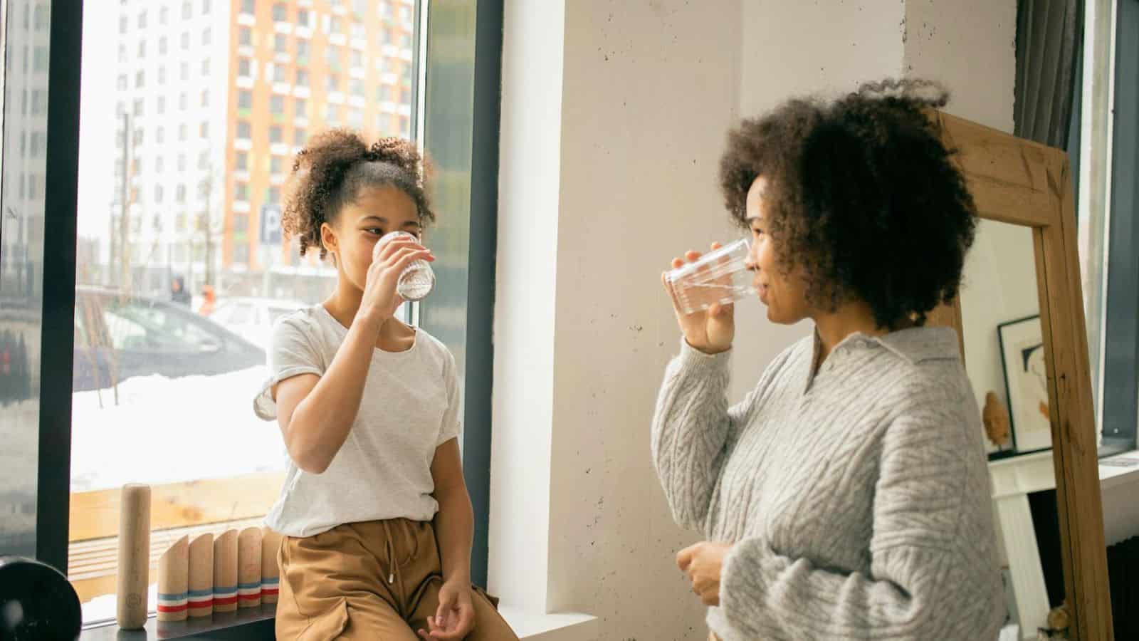 Two people with curly hair are drinking water from clear glasses indoors near a large window. One is sitting on a windowsill, and the other is standing. They appear to be in a cozy room with modern decor, including a wooden mirror and some toys on the sill.