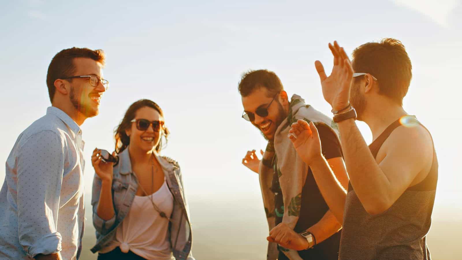 Four people are standing outside, smiling and laughing. Three men and one woman are casually dressed and wearing sunglasses. The sky is clear, suggesting a sunny day. They appear to be in good spirits, enjoying the moment together.