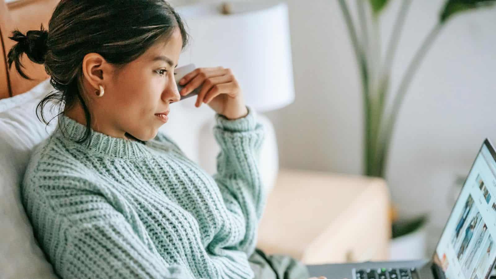 A woman wearing a light green sweater is sitting on a bed, looking at a laptop in front of her. She is resting her head on her hand, appearing focused. There is a bedside table with a lamp in the background.