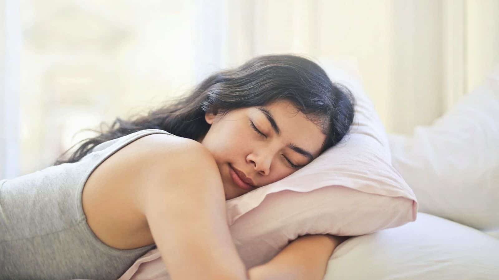 A person with long dark hair is lying on their side, eyes closed, and appears to be sleeping on a pillow with a light pink pillowcase. They are wearing a gray tank top and are positioned on a bed with a white sheet in a softly lit room.