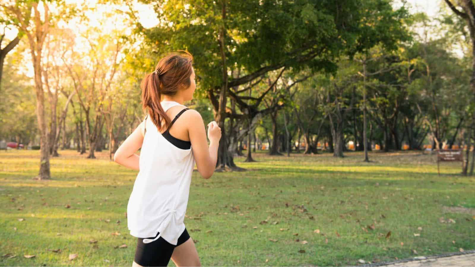A person with a ponytail, dressed in a white tank top and black shorts, is jogging on a path in a park. Trees and grassy areas are visible, with sunlight filtering through the leaves.