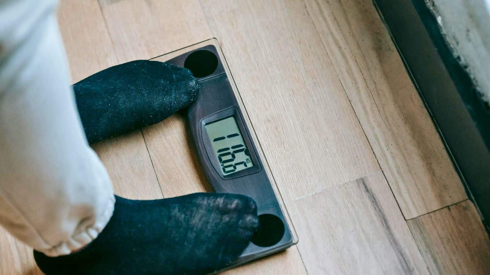 A person wearing black socks stands on a digital bathroom scale displaying a weight of 106.8 pounds. The scale is placed on a wooden floor next to a wall.