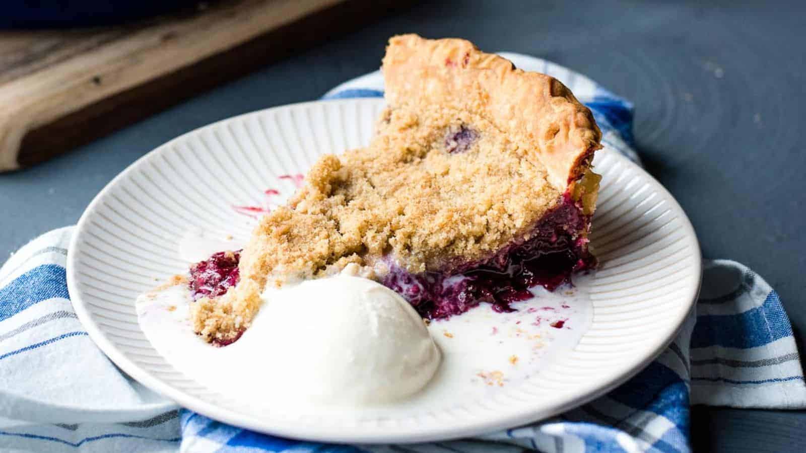 A slice of berry crumble pie is served on a white plate with a scoop of vanilla ice cream, epitomizing fall comfort food. The plate sits on a blue and white striped napkin, and the filling appears to be leaking onto the plate. The background includes a wooden cutting board.