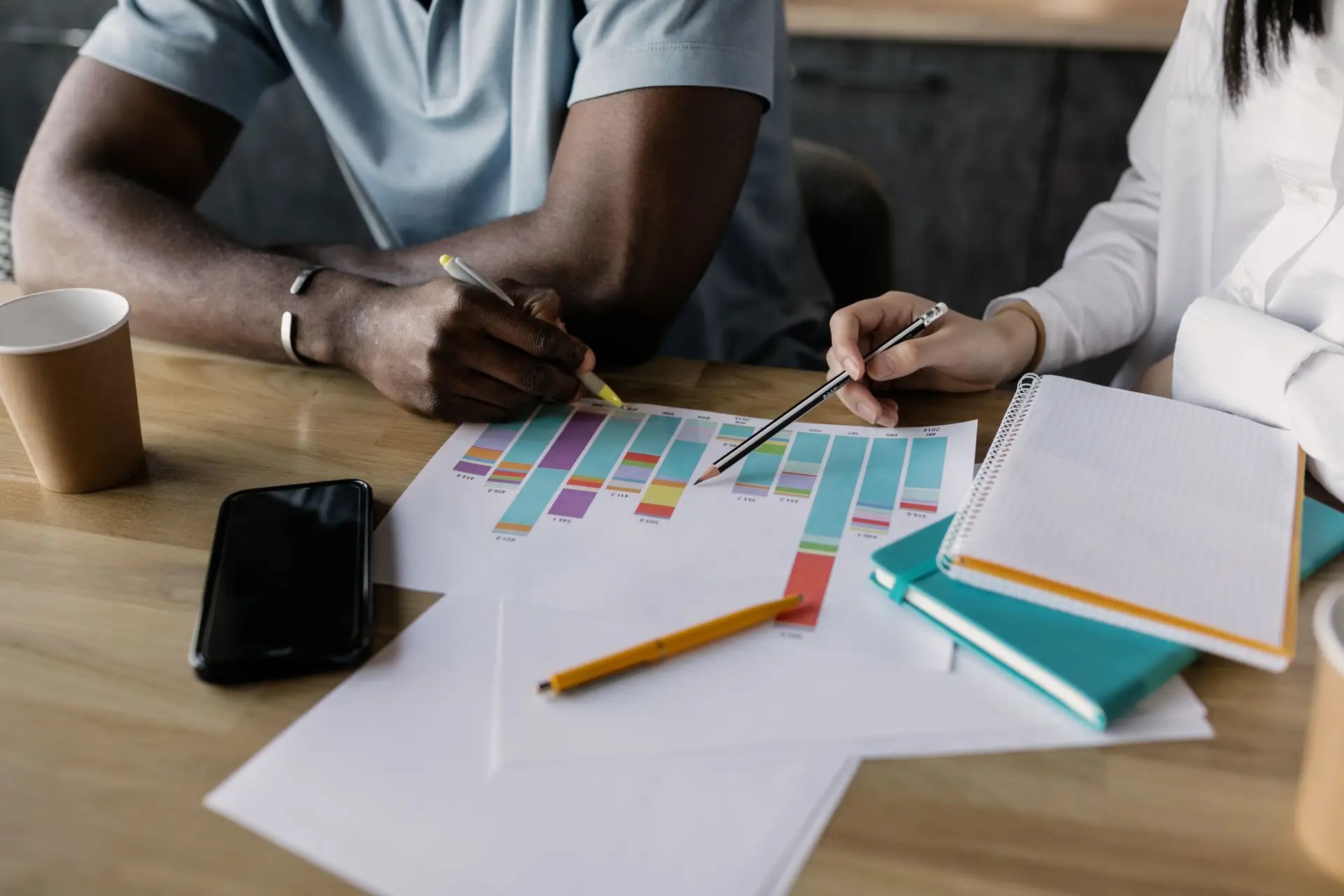 Two people review and compare data in a colorful bar chart, pointing to different results with pencils at a desk. Courtesy of Pexels