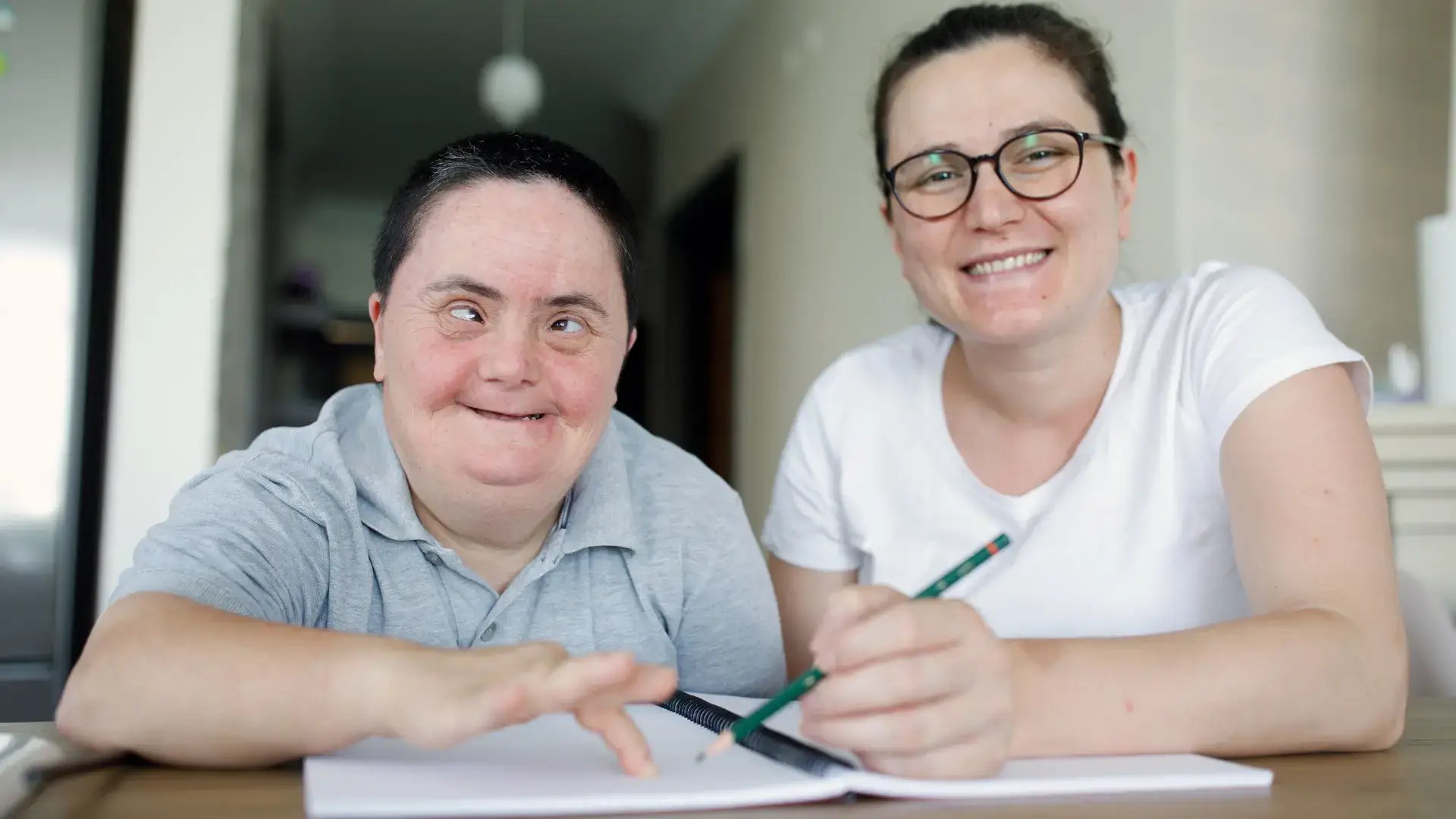 A smiling man with Down syndrome and a smiling woman sit together at a table with an open notebook and pencil.