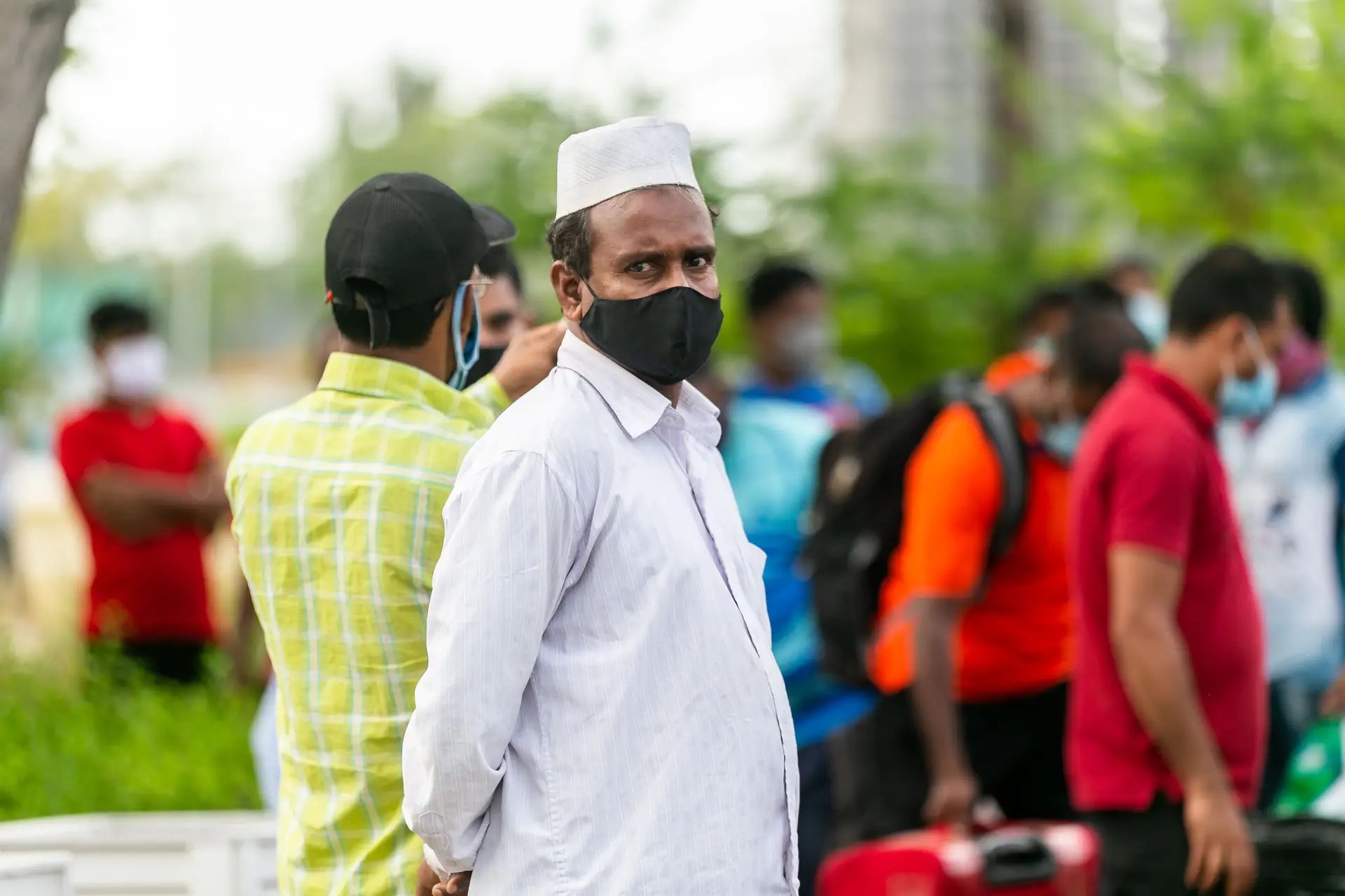 A man in India wearing a white topi and black face mask stands among a group of people outdoors. Courtesy of Pexels.
