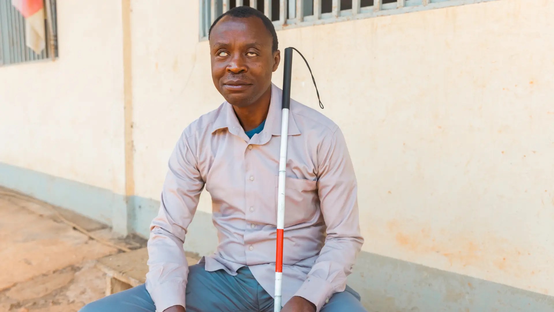 A blind man sits outdoors beside a building, holding a white cane with a red stripe upright against his shoulder. Courtesy of Canva Pro.