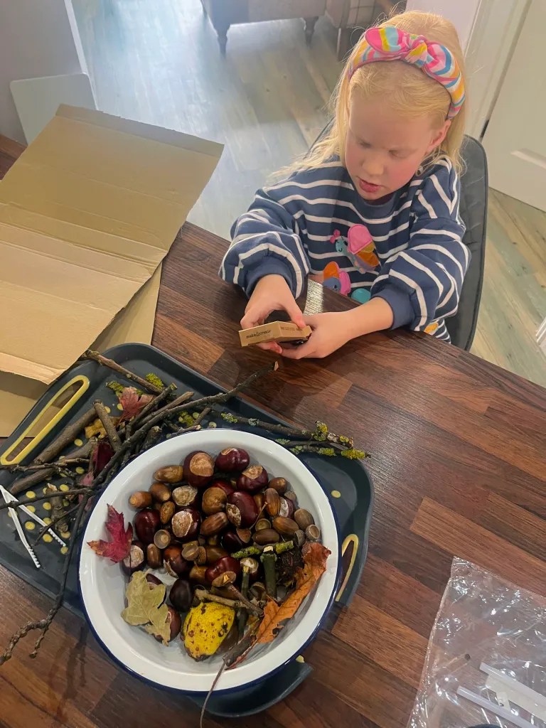 Creating Magical Autumn Nature People: A Complete EYFS Activity Guide 3 A child sitting at a table, engaged in crafting activities with natural materials such as leaves, twigs, and chestnuts displayed in a bowl.