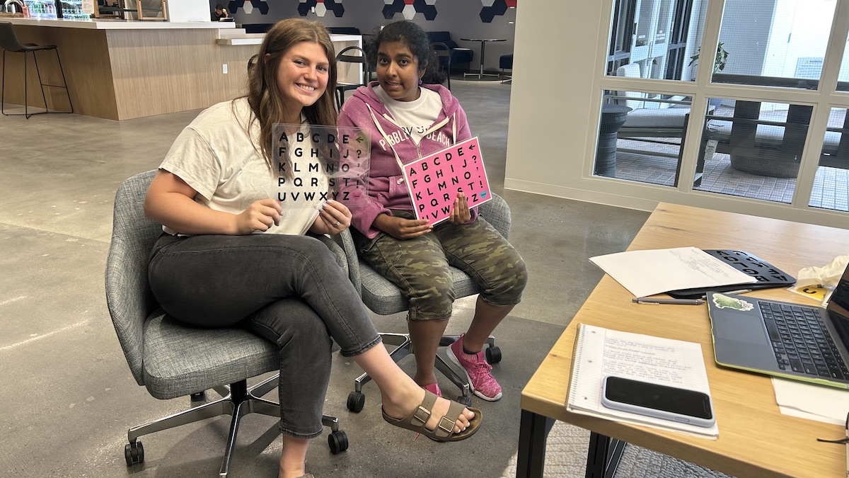 This image shows Siri (right) sitting with a letter board in her hand, with her communication coach, Kinsey Showers, sitting next to her at her home in Burlingame. (Photo: Ashwini Gangal)
