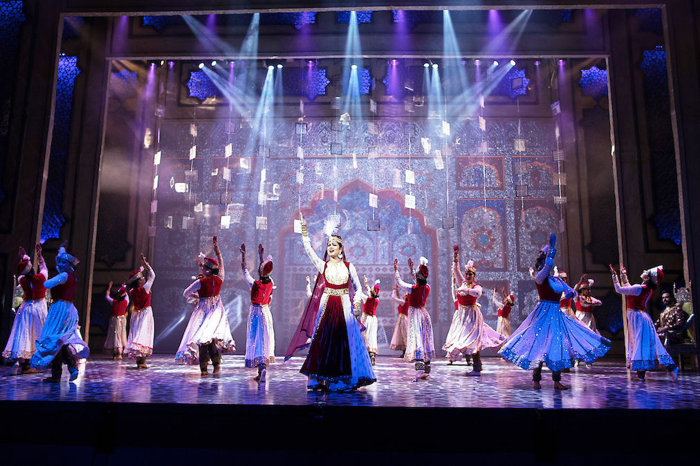 This is a scene from Mughal-e-Azam: The Musical. It shows a woman dressed in a red-and-white anarkali suite, singing, with Kathak dancers around her.(Image courtesy: MEA- DEEPESH)
