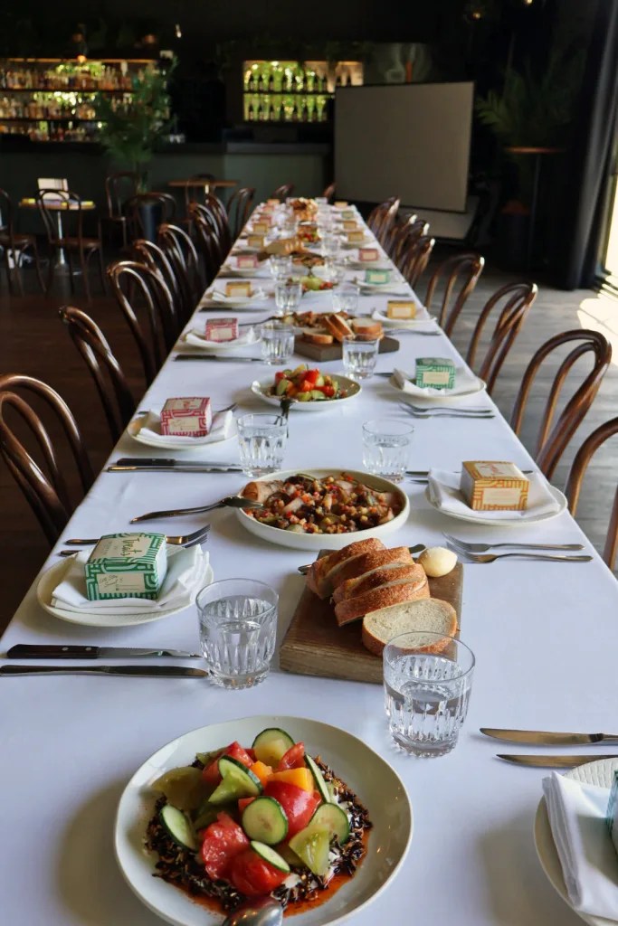 A long dining table set with plates of salad, bread, glassware, cutlery, and small boxes awaits a group meal in a restaurant with wooden chairs—an ambience as refined as Huntsman tailoring.