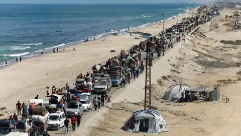 Reuters Image showing a road running next to a sandy beach, showing the sea. The road is full of grid-locked cars stretching into the distance, many with luggage and belongings strapped to their roofs. There are people walking too. On the right-hand side are tents on the right-hand side.
