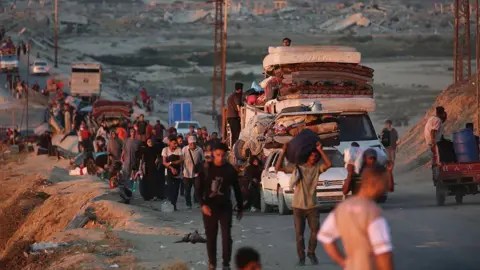 Anadolu via Getty Images Dozens of people carrying heavy luggage, using cars and carts, walk down a barren road at dusk.