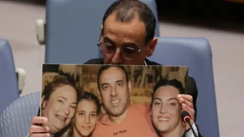 Reuters Eli Sharabi, a former Israeli hostage released by Hamas in Gaza last month, holds of a photograph of his wife and two daughters killed by Hamas, as he addresses a meeting of the United Nations Security Council