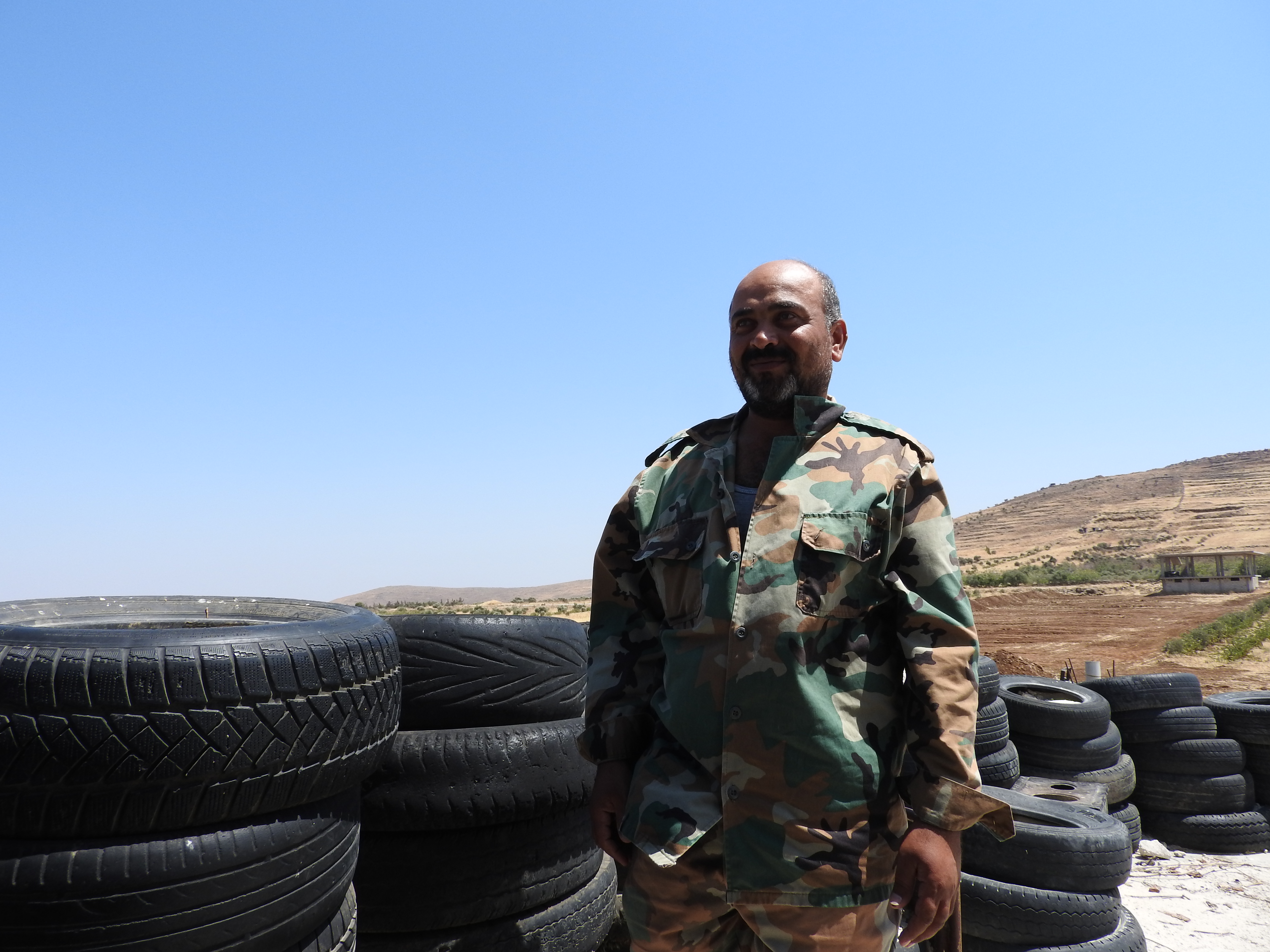 In August 2016, I went to the village of Ta’aouna, I met with residents of neighbouring Aqrab, which in December 2012 was attacked by the so-called “Free Syrian Army” who massacred between 120-150 Aqrab residents. Standing on the roof of the home to which three Aqrab survivors had come to give their testimonies, the village of Aqrab, roughly 500 metres away, was distinctly visible—as are any people in Ta’aouna who go rooftop (for laundry, water or other reasons) to terrorist snipers in the hills near Aqrab. The home owner pointed out holes from such snipers’ bullets prior. Two hundred metres down a lane, some fifteen houses remain inhabited by local Ta’aouna families (including children), in homes 300 metres from where terrorists and their snipers lie.  When terrorists massacred villagers in Aqrab in December 2012, they were then known as “Free Syrian Army” terrorists.  Now, occupied villages in the region comprise terrorists from Jabhat al-Nusra, Ahrar al-Sham, Jaysh al-Islam, and Da’esh (ISIS). As most Syrians I’ve met say, they are the same, with different names and financial backers, but commit the same heinous beheadings, assassinations, kidnappings and other western-sanctioned crimes in Syria. Rooftop the home closest to the dirt embankment beyond (this particular house uninhabited, although only 5 metres from the next inhabited one), Abu Abdo, a local defense volunteer explains how he and others in the village take night shifts to watch for attempted terrorist infiltrations. The Syrian Arab Army has hilltop posts around Ta’aouna, but nonetheless the village defenders (including many who are family men and formerly served in the SAA) watch to see if/where terrorists are shooting from/at. “We organized ourselves, since 2011. We communicate with the army and give them targets, and they do the same with us,” he says of the watch for terrorist attacks. We sit behind a wall of tires, some concrete blocks to one side serving as a defensive wall from behind which to watch for and shoot at terrorists. A second local defender appears, greets me with a friendly handshake, explains that in late 2013 terrorists managed to advance to the low hills to our right. But not since. I ask Abu Abdo what he did prior to the war on Syria. A school principal, and he still is, he does the defense volunteering after hours. I ask about his family. He has 5 kids, including the pretty 9th grade girl with long curly hair who had served us coffee and cold water in the home where Aqrab residents gave their testimonies. “I’m a principal. I used to teach in Raqqa years ago. We all got along, were peaceful. I’d walk for many kilometres in areas I wasn’t familiar with but I never had problems. I also taught in Aqrab for two years.” They point to the land between Ta’aouna and the low hills flanking the village, and the start of Aqrab beyond. “That small cement building on the land, right near there, about one month ago, a university student was shot in his head and killed, by a terrorist sniper. He was an engineering student.” Earlier they’d told me about this, and about another university student who roughly 2 weeks ago was torn apart by shelling from terrorists in Aqrab. “He had just finished his exams,” they had said. Descending from the roof, we walk past a nearby house, the children on the porch stoop. The second defense soldier tells me, with a proud smile, they are his kids. He takes me to the side of the house to show three creatively covered holes, “Dushkie” shots from the terrorists about 10 days ago. https://ingaza.wordpress.com/2016/08/11/updates-from-on-the-ground-in-syria-june-to-august-11/