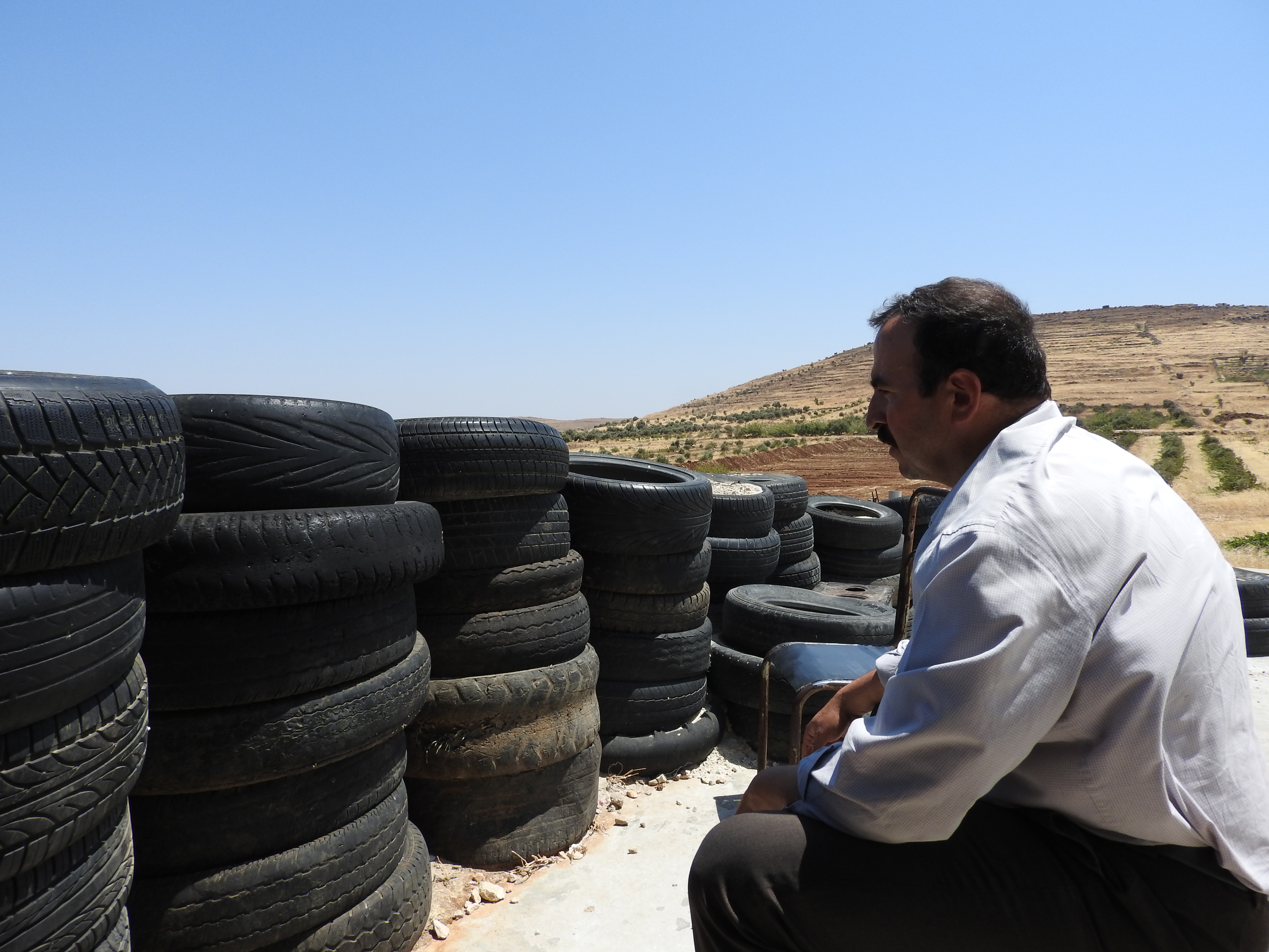 In August 2016, I went to the village of Ta’aouna, I met with residents of neighbouring Aqrab, which in December 2012 was attacked by the so-called “Free Syrian Army” who massacred between 120-150 Aqrab residents. Standing on the roof of the home to which three Aqrab survivors had come to give their testimonies, the village of Aqrab, roughly 500 metres away, was distinctly visible—as are any people in Ta’aouna who go rooftop (for laundry, water or other reasons) to terrorist snipers in the hills near Aqrab. The home owner pointed out holes from such snipers’ bullets prior. Two hundred metres down a lane, some fifteen houses remain inhabited by local Ta’aouna families (including children), in homes 300 metres from where terrorists and their snipers lie. When terrorists massacred villagers in Aqrab in December 2012, they were then known as “Free Syrian Army” terrorists. Now, occupied villages in the region comprise terrorists from Jabhat al-Nusra, Ahrar al-Sham, Jaysh al-Islam, and Da’esh (ISIS). As most Syrians I’ve met say, they are the same, with different names and financial backers, but commit the same heinous beheadings, assassinations, kidnappings and other western-sanctioned crimes in Syria. Rooftop the home closest to the dirt embankment beyond (this particular house uninhabited, although only 5 metres from the next inhabited one), Abu Abdo, a local defense volunteer explains how he and others in the village take night shifts to watch for attempted terrorist infiltrations. The Syrian Arab Army has hilltop posts around Ta’aouna, but nonetheless the village defenders (including many who are family men and formerly served in the SAA) watch to see if/where terrorists are shooting from/at. “We organized ourselves, since 2011. We communicate with the army and give them targets, and they do the same with us,” he says of the watch for terrorist attacks. We sit behind a wall of tires, some concrete blocks to one side serving as a defensive wall from behind which to watch for and shoot at terrorists. A second local defender appears, greets me with a friendly handshake, explains that in late 2013 terrorists managed to advance to the low hills to our right. But not since. I ask Abu Abdo what he did prior to the war on Syria. A school principal, and he still is, he does the defense volunteering after hours. I ask about his family. He has 5 kids, including the pretty 9th grade girl with long curly hair who had served us coffee and cold water in the home where Aqrab residents gave their testimonies. “I’m a principal. I used to teach in Raqqa years ago. We all got along, were peaceful. I’d walk for many kilometres in areas I wasn’t familiar with but I never had problems. I also taught in Aqrab for two years.” They point to the land between Ta’aouna and the low hills flanking the village, and the start of Aqrab beyond. “That small cement building on the land, right near there, about one month ago, a university student was shot in his head and killed, by a terrorist sniper. He was an engineering student.” Earlier they’d told me about this, and about another university student who roughly 2 weeks ago was torn apart by shelling from terrorists in Aqrab. “He had just finished his exams,” they had said. Descending from the roof, we walk past a nearby house, the children on the porch stoop. The second defense soldier tells me, with a proud smile, they are his kids. He takes me to the side of the house to show three creatively covered holes, “Dushkie” shots from the terrorists about 10 days ago. https://ingaza.wordpress.com/2016/08/11/updates-from-on-the-ground-in-syria-june-to-august-11/