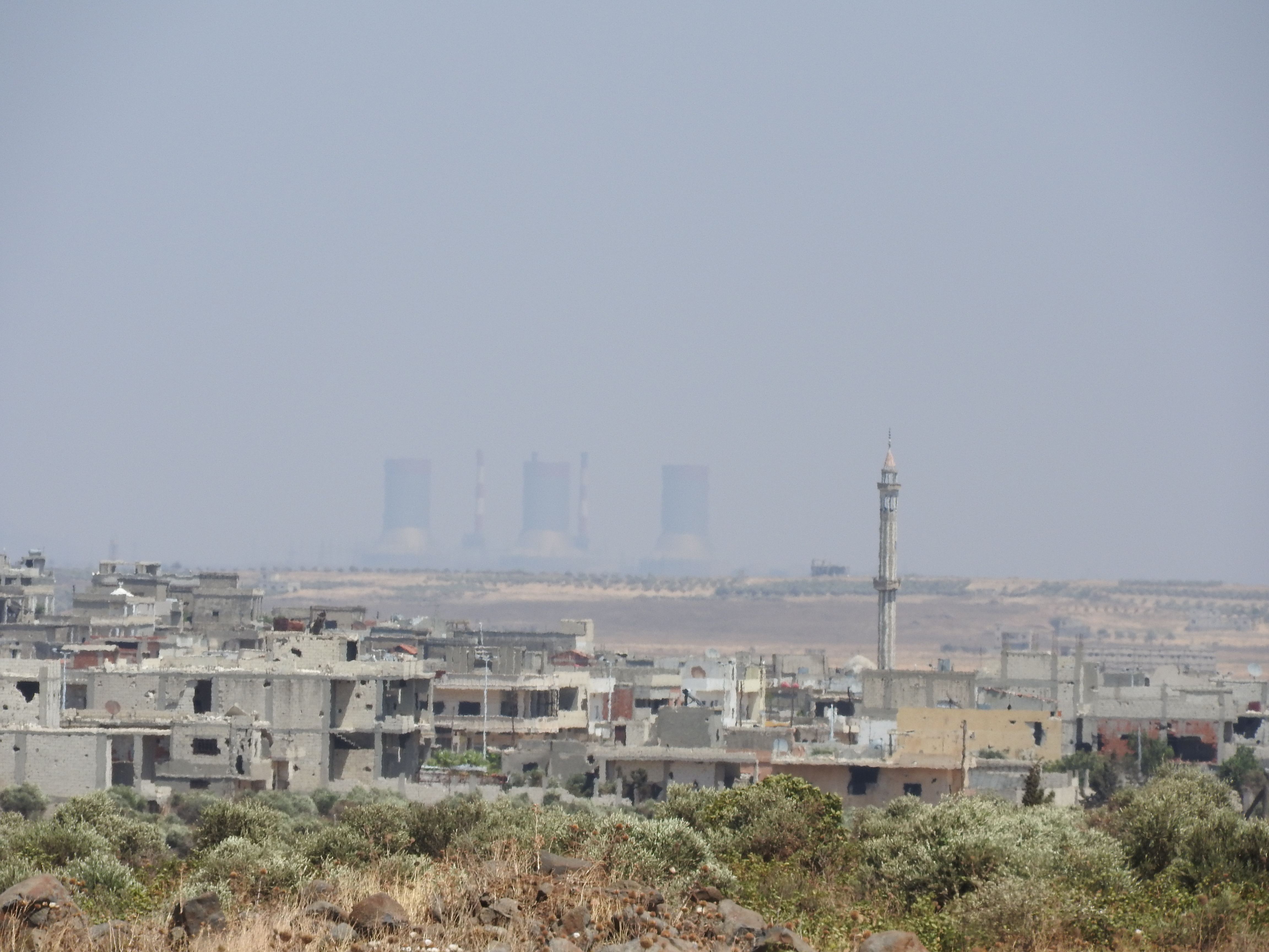 In August 2016, I went to the village of Ta’aouna, I met with residents of neighbouring Aqrab, which in December 2012 was attacked by the so-called “Free Syrian Army” who massacred between 120-150 Aqrab residents. Standing on the roof of the home to which three Aqrab survivors had come to give their testimonies, the village of Aqrab, roughly 500 metres away, was distinctly visible—as are any people in Ta’aouna who go rooftop (for laundry, water or other reasons) to terrorist snipers in the hills near Aqrab. The home owner pointed out holes from such snipers’ bullets prior. Two hundred metres down a lane, some fifteen houses remain inhabited by local Ta’aouna families (including children), in homes 300 metres from where terrorists and their snipers lie. When terrorists massacred villagers in Aqrab in December 2012, they were then known as “Free Syrian Army” terrorists. Now, occupied villages in the region comprise terrorists from Jabhat al-Nusra, Ahrar al-Sham, Jaysh al-Islam, and Da’esh (ISIS). As most Syrians I’ve met say, they are the same, with different names and financial backers, but commit the same heinous beheadings, assassinations, kidnappings and other western-sanctioned crimes in Syria. Rooftop the home closest to the dirt embankment beyond (this particular house uninhabited, although only 5 metres from the next inhabited one), Abu Abdo, a local defense volunteer explains how he and others in the village take night shifts to watch for attempted terrorist infiltrations. The Syrian Arab Army has hilltop posts around Ta’aouna, but nonetheless the village defenders (including many who are family men and formerly served in the SAA) watch to see if/where terrorists are shooting from/at. “We organized ourselves, since 2011. We communicate with the army and give them targets, and they do the same with us,” he says of the watch for terrorist attacks. We sit behind a wall of tires, some concrete blocks to one side serving as a defensive wall from behind which to watch for and shoot at terrorists. A second local defender appears, greets me with a friendly handshake, explains that in late 2013 terrorists managed to advance to the low hills to our right. But not since. I ask Abu Abdo what he did prior to the war on Syria. A school principal, and he still is, he does the defense volunteering after hours. I ask about his family. He has 5 kids, including the pretty 9th grade girl with long curly hair who had served us coffee and cold water in the home where Aqrab residents gave their testimonies. “I’m a principal. I used to teach in Raqqa years ago. We all got along, were peaceful. I’d walk for many kilometres in areas I wasn’t familiar with but I never had problems. I also taught in Aqrab for two years.” They point to the land between Ta’aouna and the low hills flanking the village, and the start of Aqrab beyond. “That small cement building on the land, right near there, about one month ago, a university student was shot in his head and killed, by a terrorist sniper. He was an engineering student.” Earlier they’d told me about this, and about another university student who roughly 2 weeks ago was torn apart by shelling from terrorists in Aqrab. “He had just finished his exams,” they had said. Descending from the roof, we walk past a nearby house, the children on the porch stoop. The second defense soldier tells me, with a proud smile, they are his kids. He takes me to the side of the house to show three creatively covered holes, “Dushkie” shots from the terrorists about 10 days ago. https://ingaza.wordpress.com/2016/08/11/updates-from-on-the-ground-in-syria-june-to-august-11/
