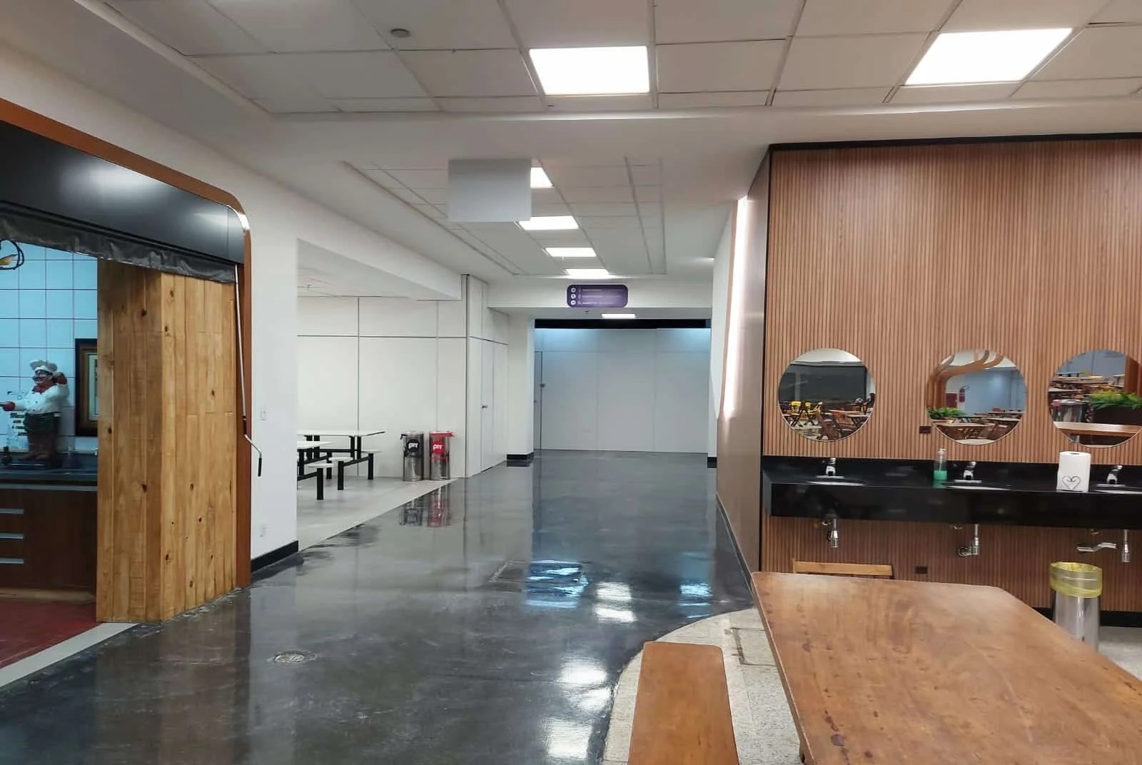 A imagem mostra um caminho com chão preto que leva aos banheiros. À esquerda, há um restaurante com uma parede de madeira e um espaço branco ao fundo, que contém uma mesa também branca. À direita, há uma parede com revestimento de madeira, com lavatórios pretos e espelhos redondos instalados acima dos lavatórios.