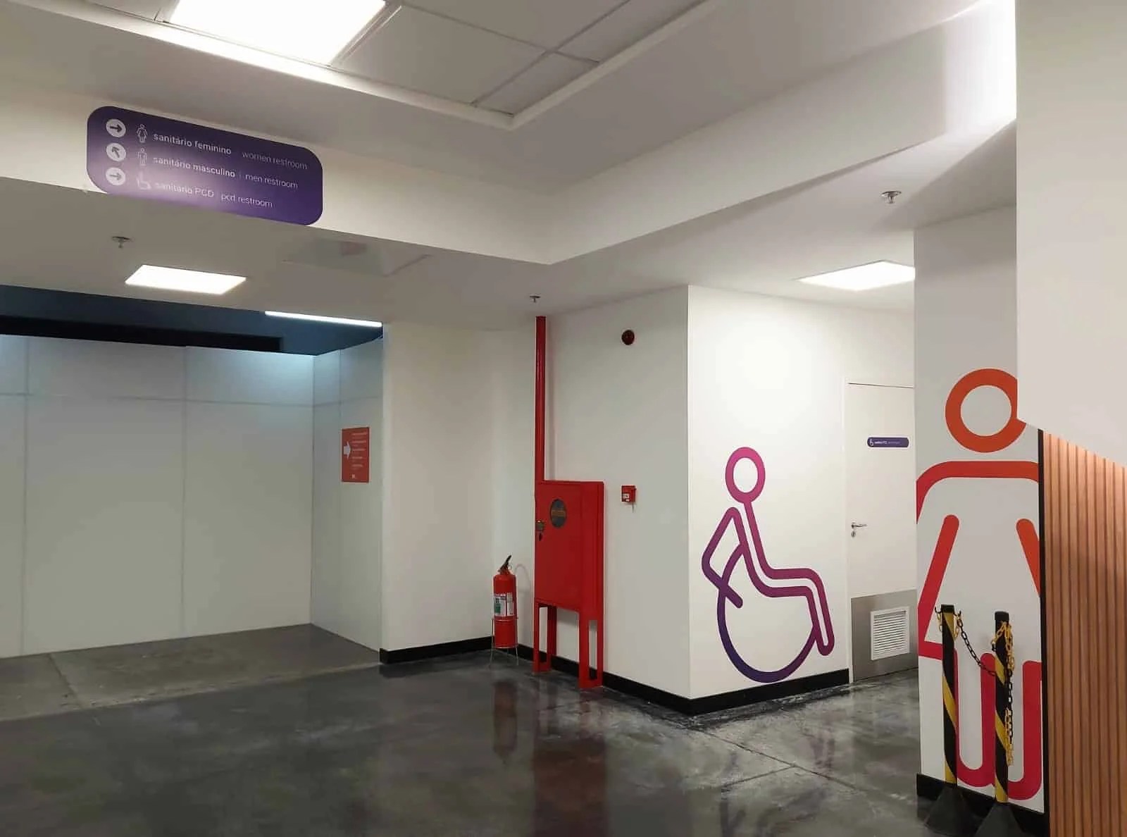 A imagem mostra a parte externa dos banheiros, estamos em frente da entrada deles. Na parte superior, próximo ao teto, há uma placa roxa com as seguintes indicações: “Sanitário feminino” com uma seta apontando para a direita, “Sanitário masculino” com uma seta diagonal para a esquerda, e “Banheiros para pessoas com deficiência” com uma seta apontando para a direita. Ao lado da placa, há uma parede branca com uma imagem em tons de roxo e rosa de uma pessoa utilizando cadeira de rodas, e a porta do banheiro acessível. Na parede à direita, há uma imagem em tons de laranja de uma mulher, indicando o sanitário feminino.