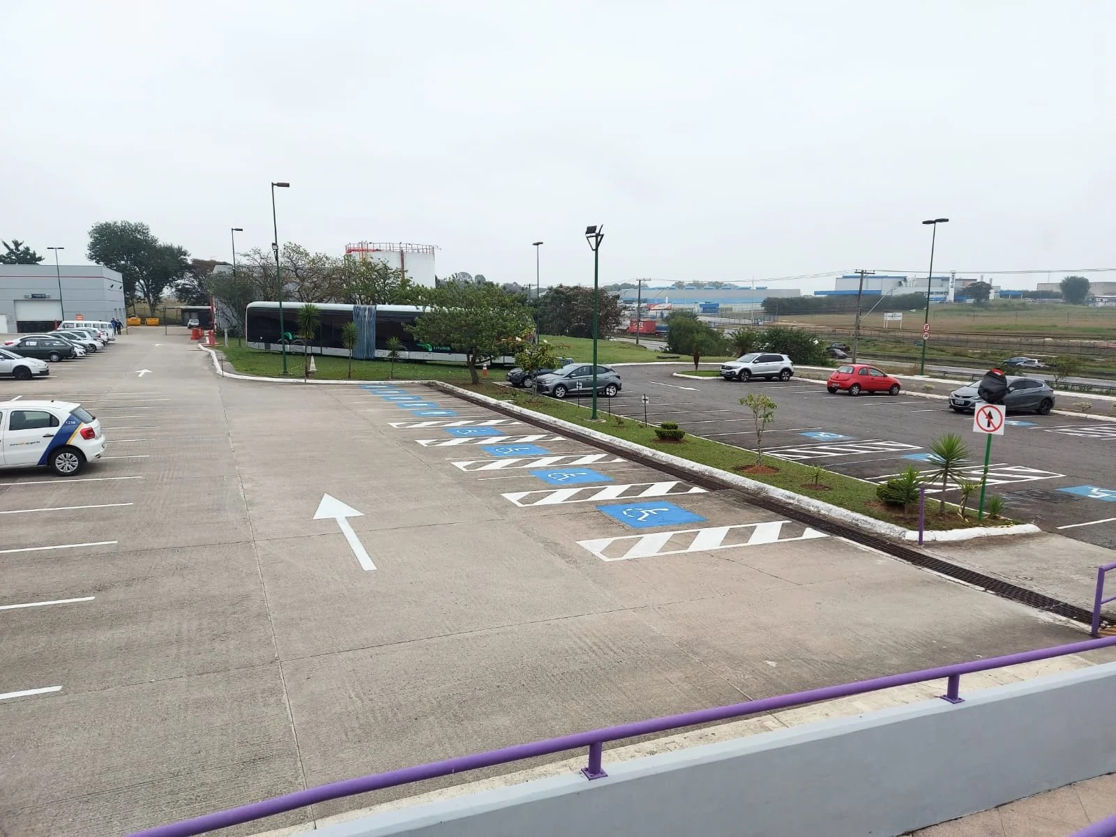 A imagem mostra um estacionamento com asfalto. Alguns carros estão estacionados nos cantos esquerdo e direito da imagem. No lado direito, há oito vagas reservadas para pessoas com deficiência. No fundo do estacionamento, um ônibus preto e cinza está estacionado sobre um gramado. O céu está nublado, indicando um dia sem sol.