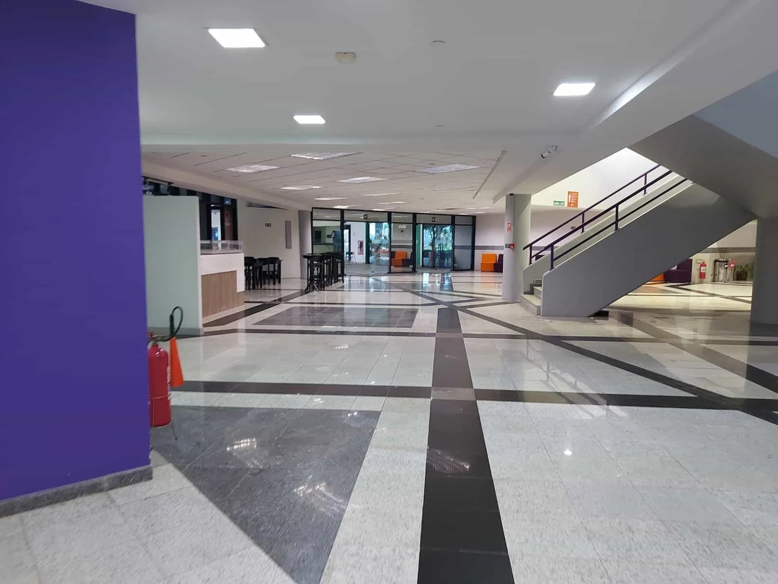 A imagem mostra o hall de eventos. Ele apresenta, à esquerda, uma parede roxa com um extintor de incêndio no chão. Atrás dessa parede há uma cafeteria branca com cadeiras pretas. No centro da imagem, o chão é branco com formas geométricas em cinza e preto. À direita, há uma escadaria cinza com corrimão roxo que leva ao piso superior. Ao fundo, uma porta de vidro dá acesso à parte externa, onde está localizada a Unesp.