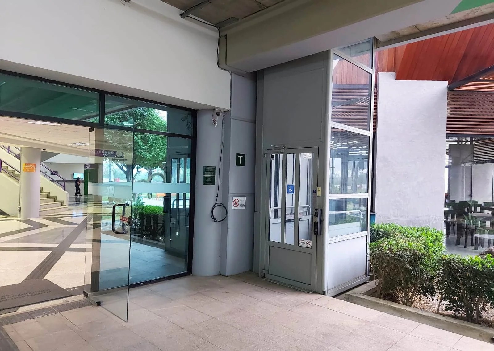 A imagem mostra uma porta de vidro que dá acesso à Área de Eventos. Ao fundo, é possível ver a escadaria. No centro da imagem, há um elevador, com pequenas plantas ao seu lado, ele está no térreo, por isso uma placa na parede traz a letra “T”.
