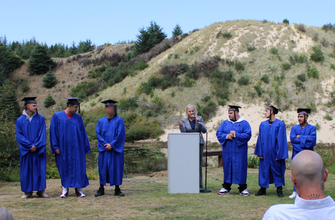 three graduates in blue cap and gown standing outside with principal at a podium