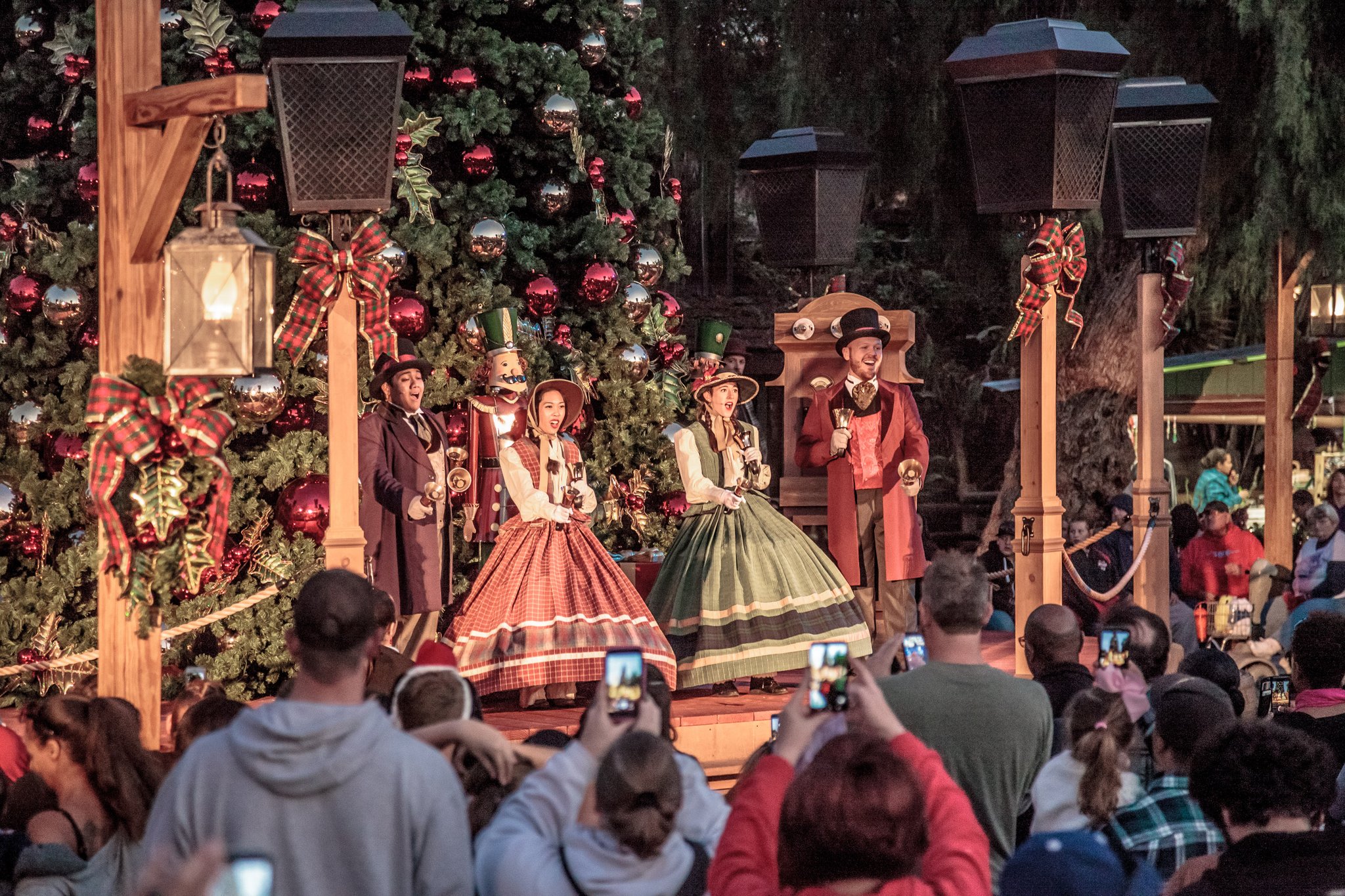 Carolers sing under the night sky with Christmas decor surrounding.