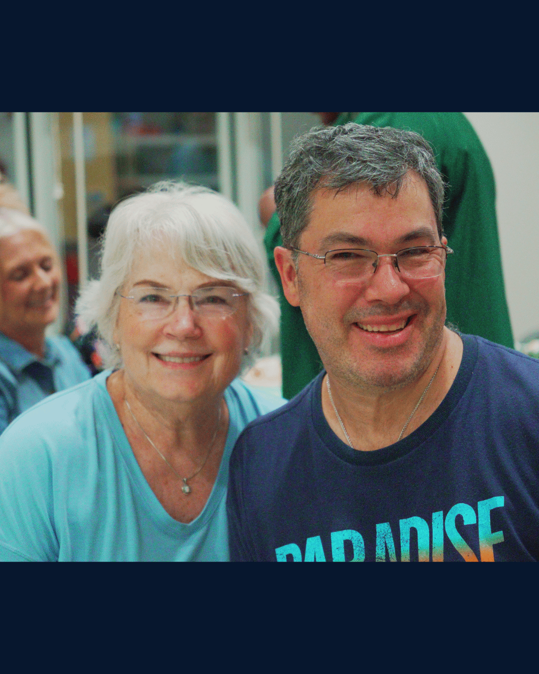 An older woman with white hair and glasses and a man with short gray hair and glasses smile warmly together at a Social Circle of Life dance event, both dressed in teal and navy.