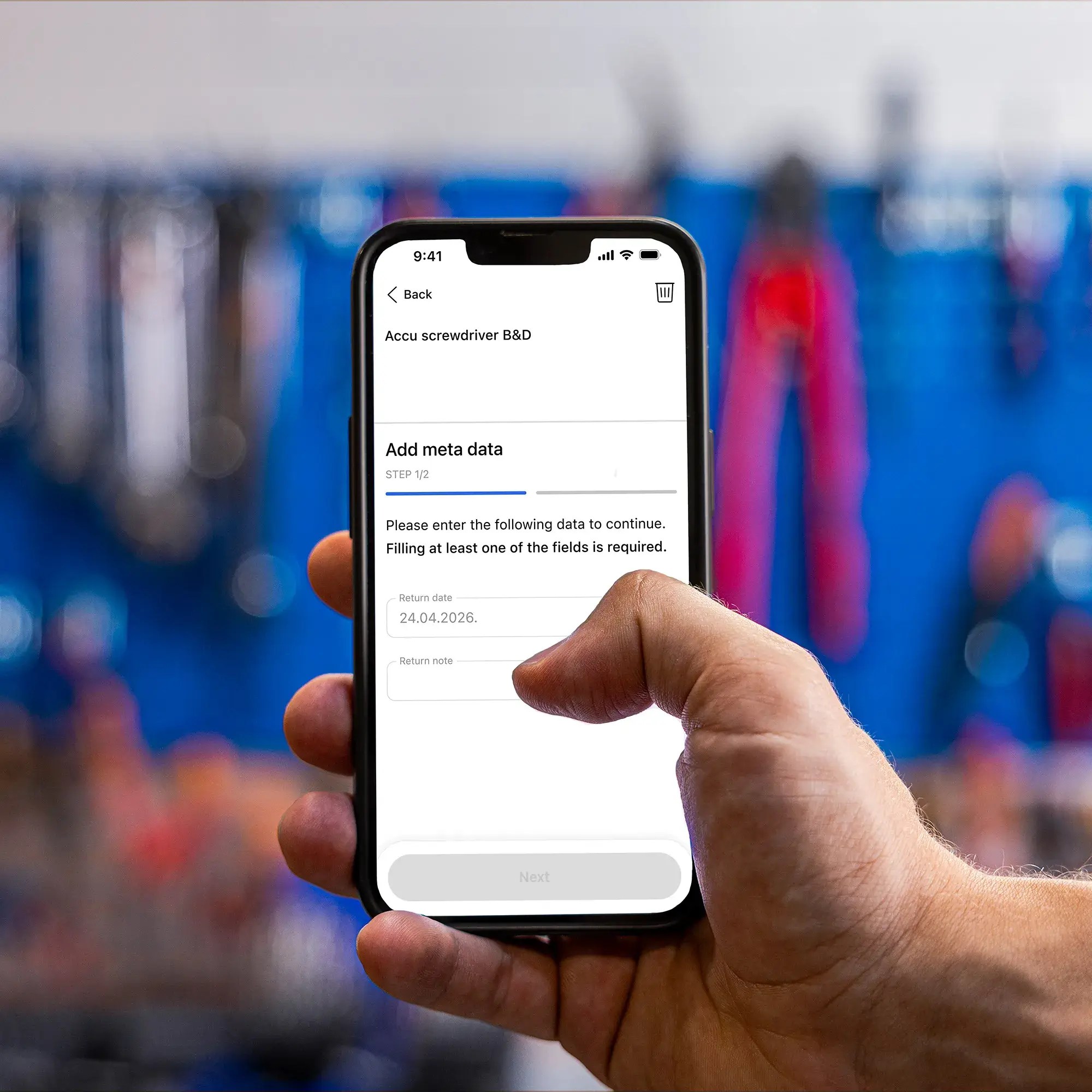 Hand holding a smartphone with the Invendor Trace app, adding a return date for a borrowed screwdriver, with a tool wall in the background.