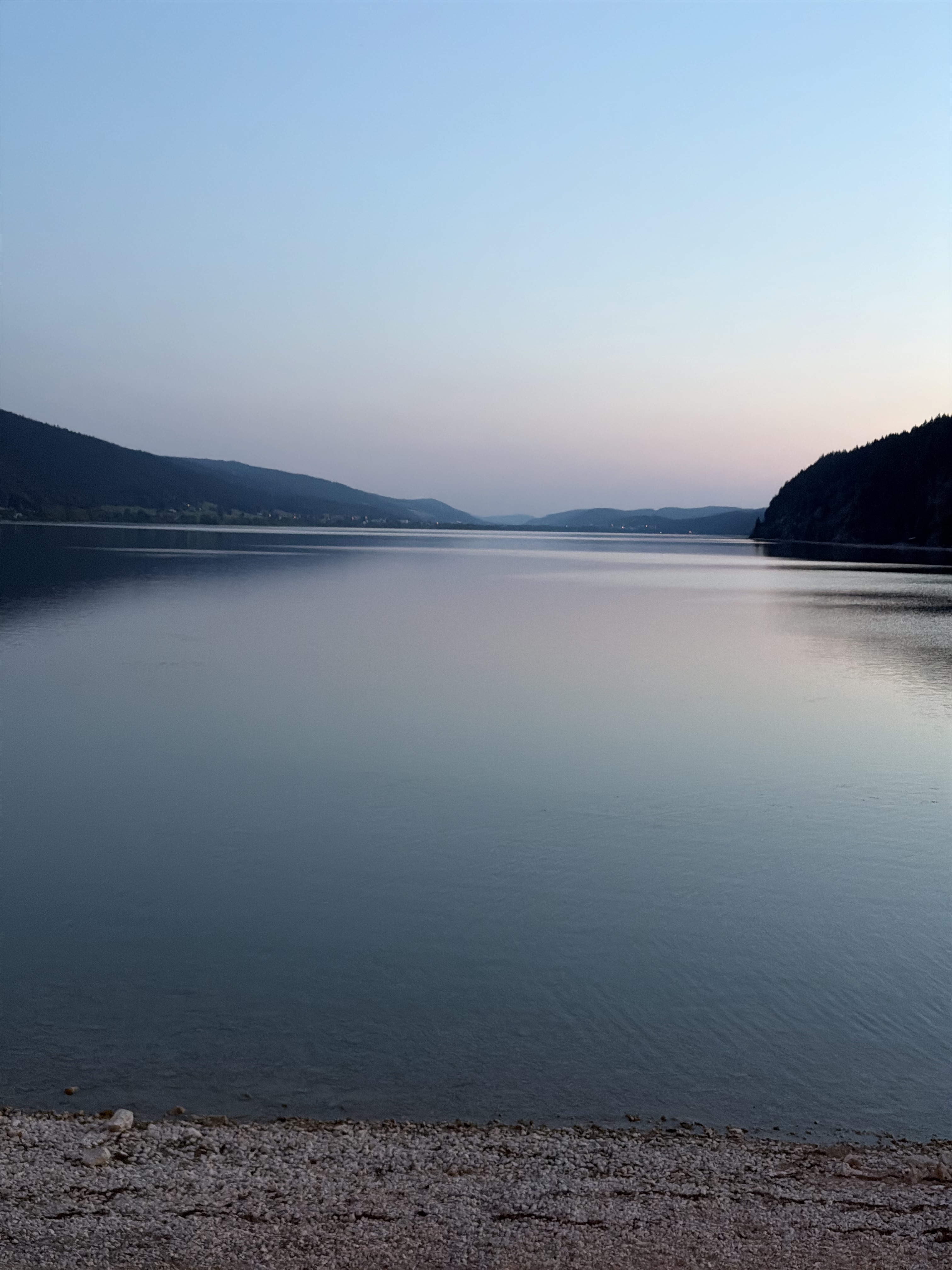 Calm water of the Lac de Joux at dusk, ringed by dark forested hills under a pale pink and blue sky