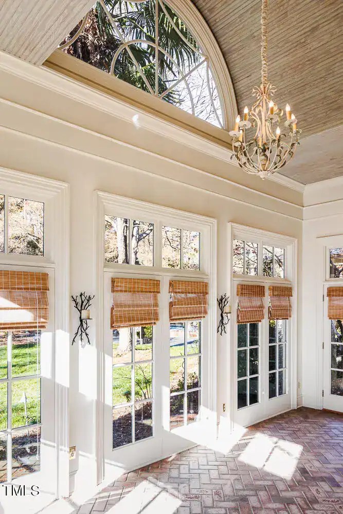Bright sunlit interior with large windows, white trim, and bamboo blinds, showcasing elegant chandeliers and classic architecture, perfect for a sophisticated floral design setting or home decor inspiration.