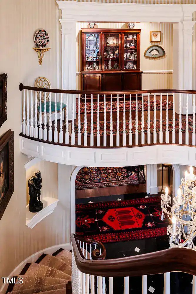 Elegant Victorian-style interior featuring a grand staircase with white balustrades and rich red and black oriental rugs, complemented by antique furniture and classic decor details.