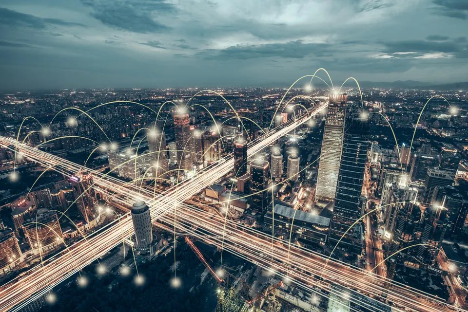 Nighttime city skyline with glowing arcs of light linking skyscrapers to illustrate a connected smart city network. Between-buildings light trails suggest data flow and connectivity.